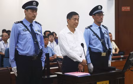 Bạc Hy Lai đứng giữa hai cảnh sát cao lớn, và ám hiệu khó hiểu ở tay trái.