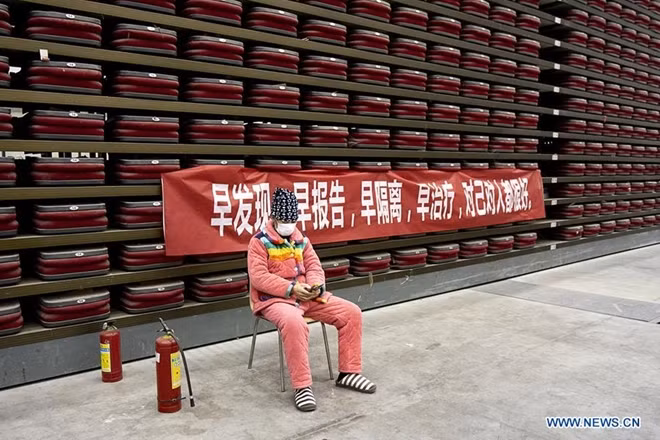 Một bệnh nhân ngồi sử dụng điện thoại "giết thời gian" trong thời gian tại bệnh viện, đằng sau là băng rôn với dòng chứ "phát hiện sớm, báo cáo sớm, cách ly sớm, điều trị sớm, tốt cho bản thân mọi người".