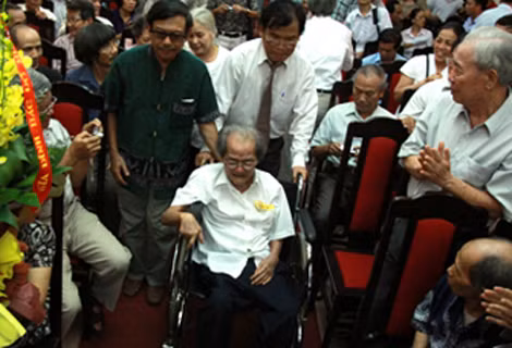 Nhà văn Sơn Tùng xúc động ngồi trên xe lăn đi vào hội trường nhận Huân chương anh hùng lao động trong sự chào đón và chúc mừng của nhiều bạn bè, đồng nghiệp.