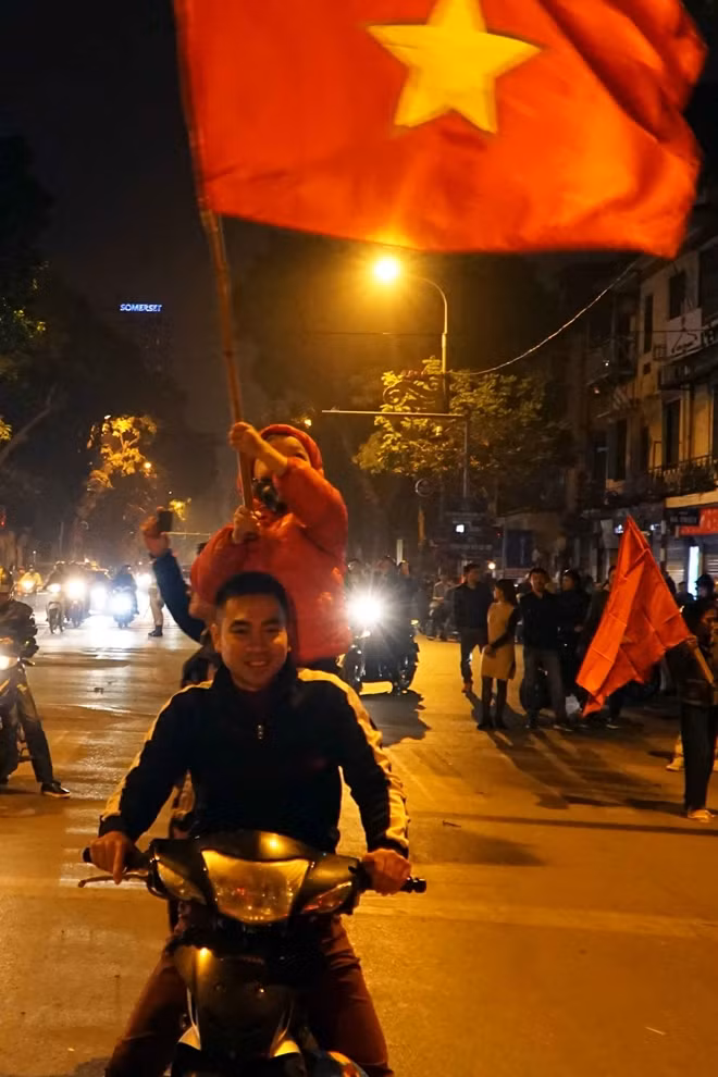 Bé trai tay cầm cờ được bố mẹ cho đứng lên xe ăn mừng.